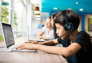 A side view of 2 children sat at a desk with a laptop in front of them. They are both wearing black headsets with blue accents on.