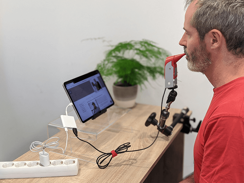 A gentleman wearing a red T-Shirt. Sat in front of him is a wooden desk on which a tablet is sat on a stand. In front of the gentleman is arm on which a mouth based mouse with a sip and puff switch