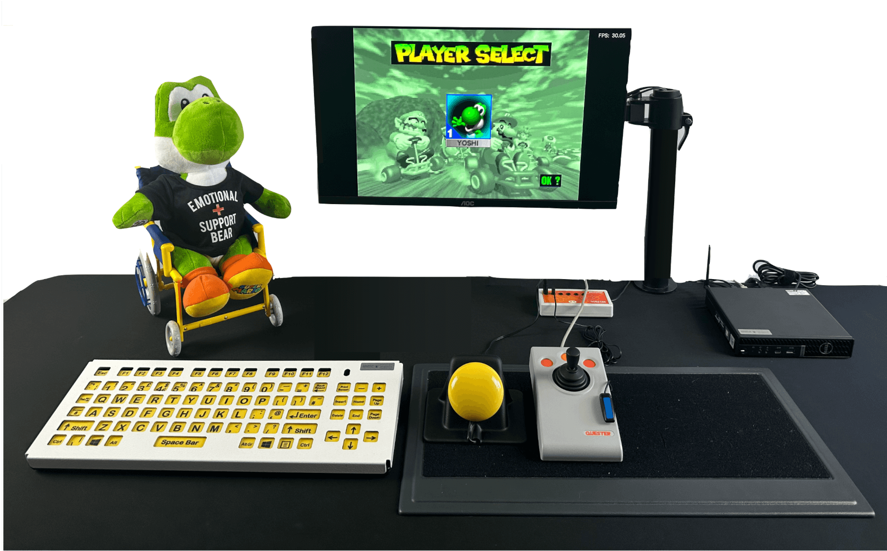 A computer setup on a black table featuring a yellow large key keyboard, a yellow assistive switch, grey joystick and an orange switch. There is a monitor mounted to the table with a Yoshi plush in a teddy wheelchair next to it.