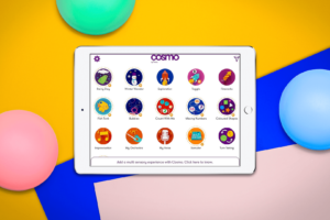 A white iPad with the Cosmo app open. It is showing 15 of the different games you can play with Cosmo. The background is yellow with blue and pink squares rotated in the bottom right corner. There a 3 Cosmo switches around the iPad, one is red, one is green and the other is blue.