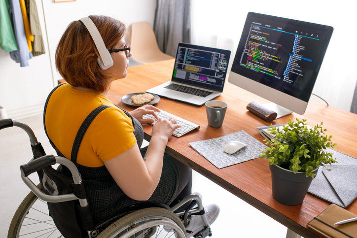Woman wearing a mustard top, sat in a manual wheelchair sat in front of a desk with a monitor and laptop, she is wearing headphones.