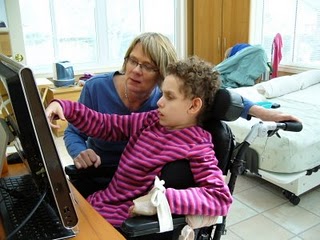 Young girl in a motorised chair, there is a lady crouched to her left helping her use assistive technology.