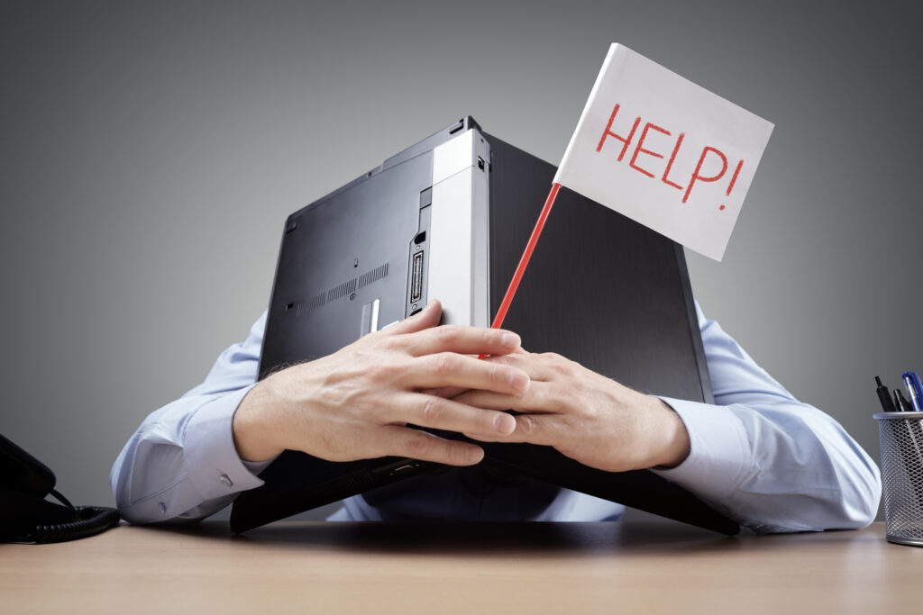 A person sat at a desk holding a laptop open like a book against his head. in his hand he is holding a flag that has Help in red on it.